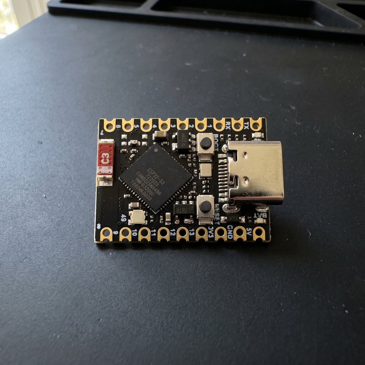 ESP32-S3 Supermini Development Kit placed on a workbench, showing the compact development board with USB Type-C connector, ESP32-S3 chip, onboard buttons, and gold-plated pin headers in a real-world workspace.