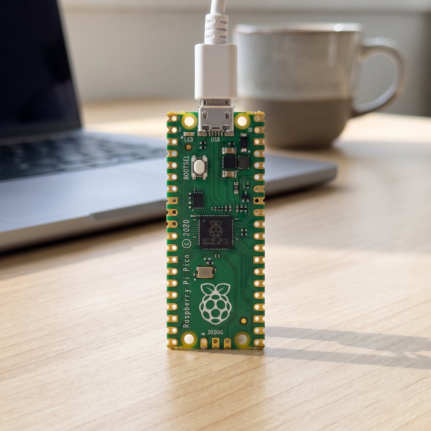 Raspberry Pi Pico board connected by USB cable on a desk with laptop in the background