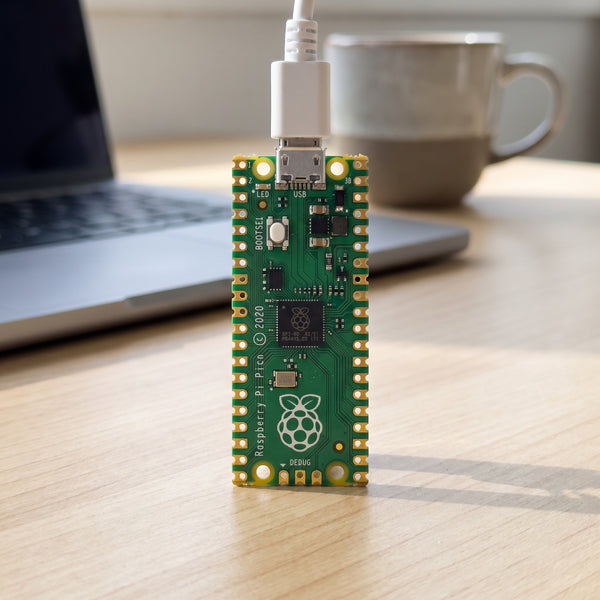 Raspberry Pi Pico board connected by USB cable on a desk with laptop in the background